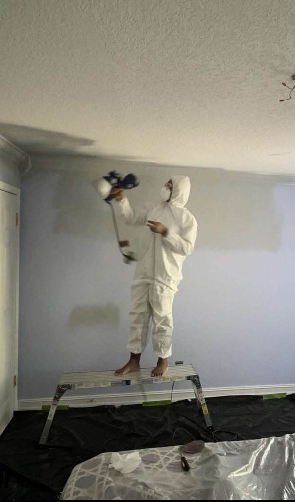Image of afro and reno in a paint suit, using a paint sprayer to prime the walls and ceiling
